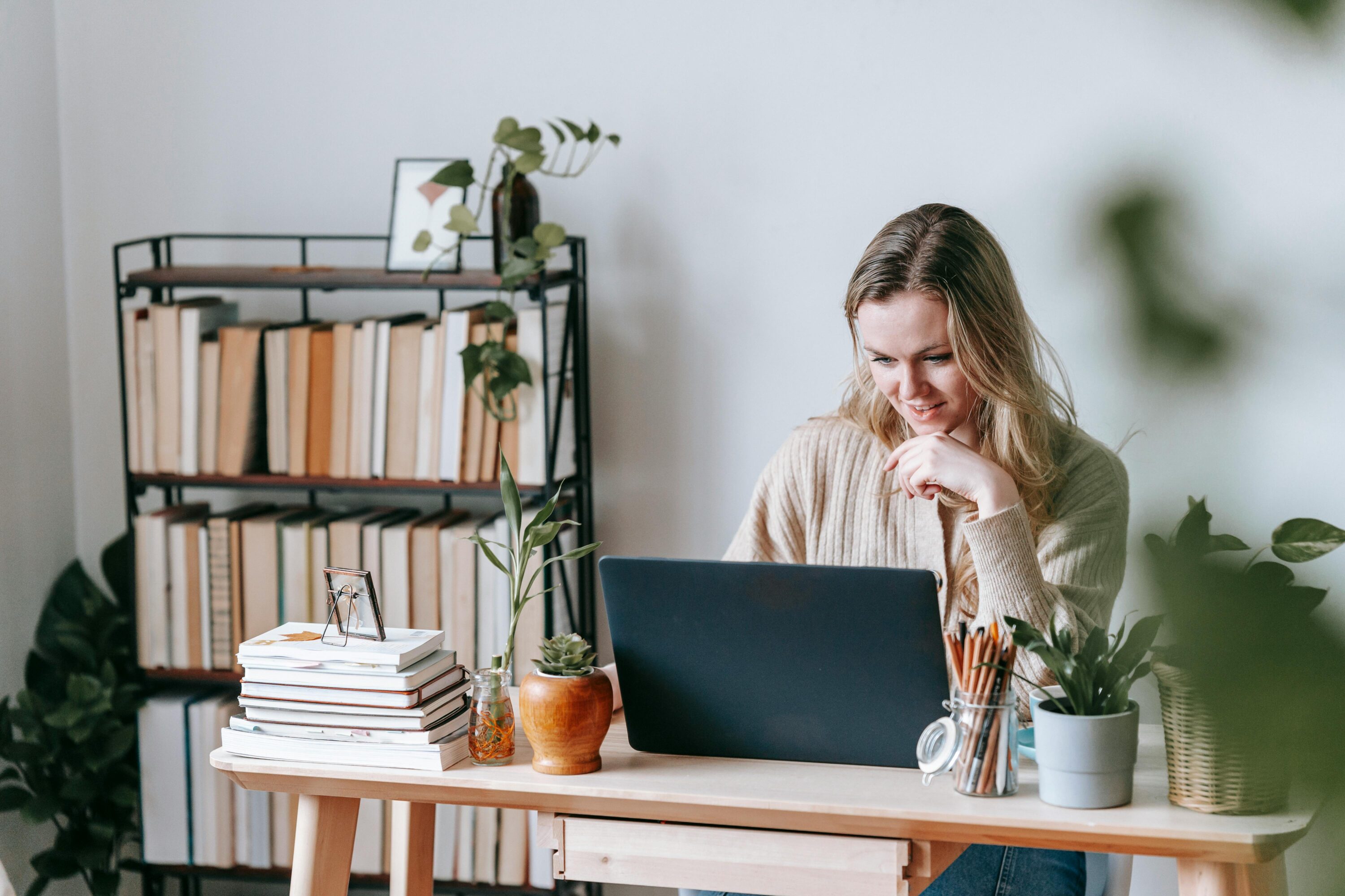 Woman in home office filled with plants, smiling at her laptop.