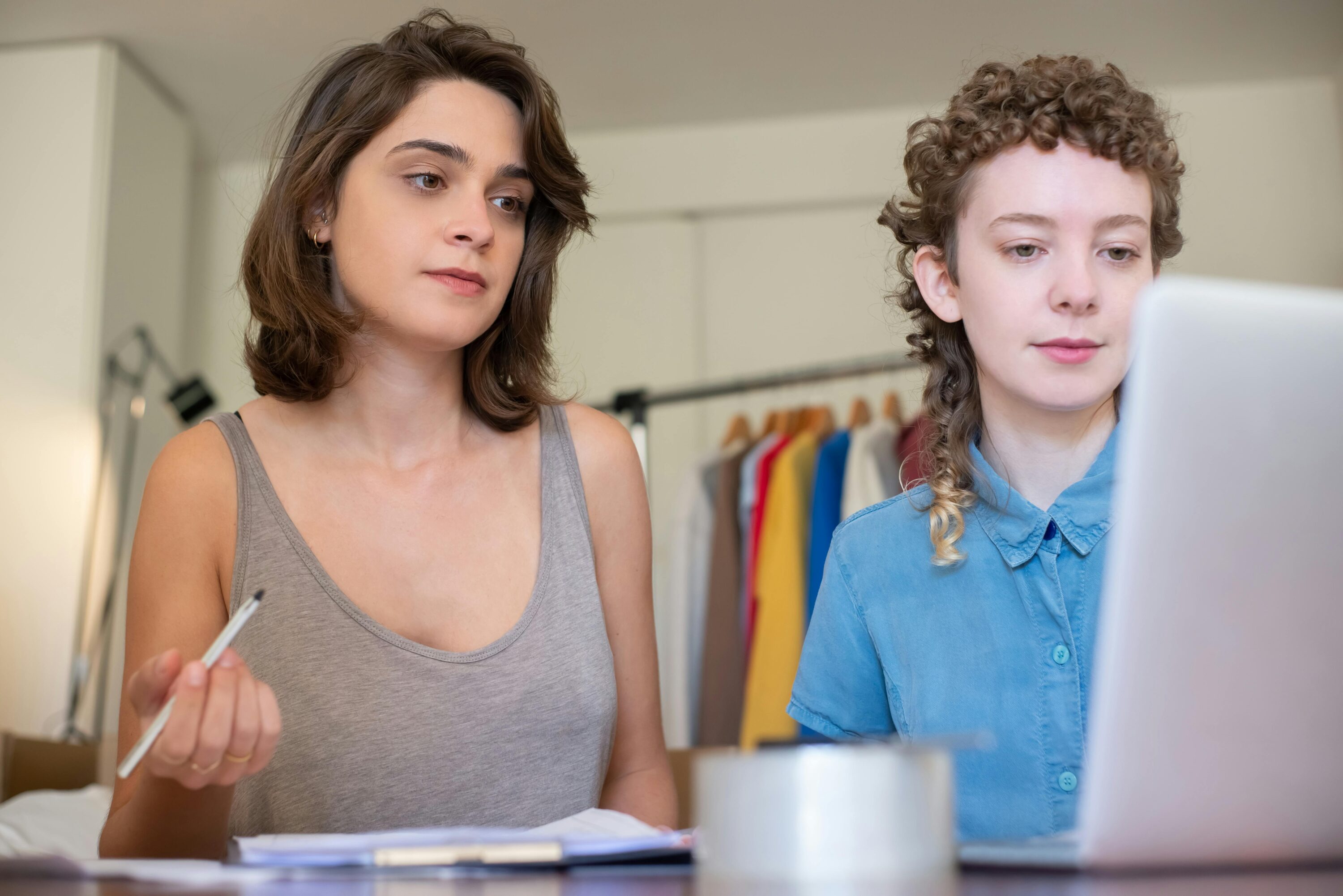 Two women looking on a laptop, with a clothing rail in the background, both brainstorming ideas.